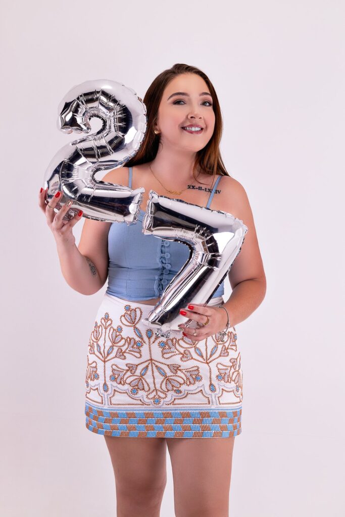 Mulher sorrindo em ensaio de aniversário, segurando balões prateados com o número 27.