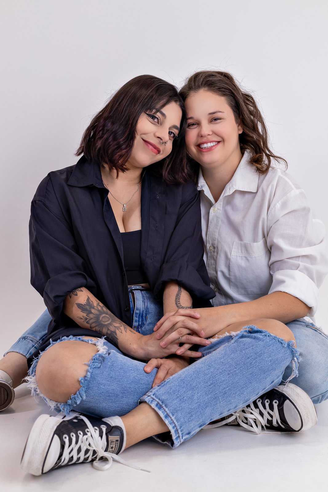 Duas mulheres sentadas juntas no estúdio, sorrindo e de mãos dadas.