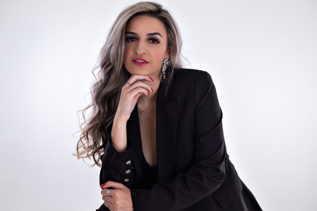 Retrato profissional de mulher com blazer preto e a mão apoiada sob o queixo, em fundo claro.