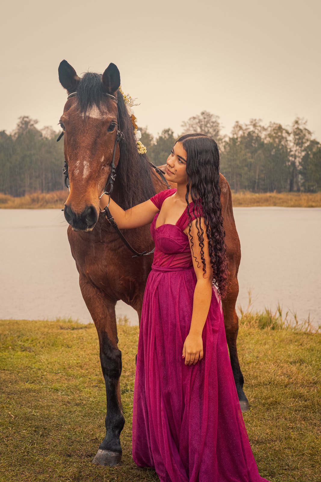 Mulher de vestido magenta ao lado de um cavalo em ensaio ao ar livre, perto de um lago.