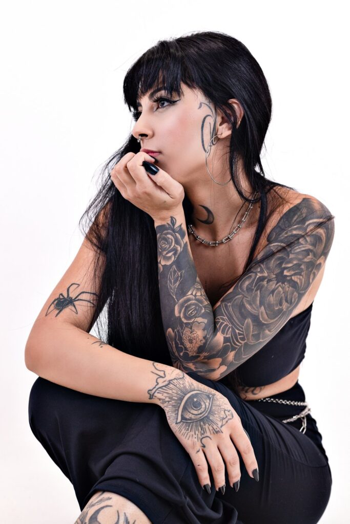 Mulher tatuada com cabelo preto, sentada em retrato de estúdio sobre fundo claro.