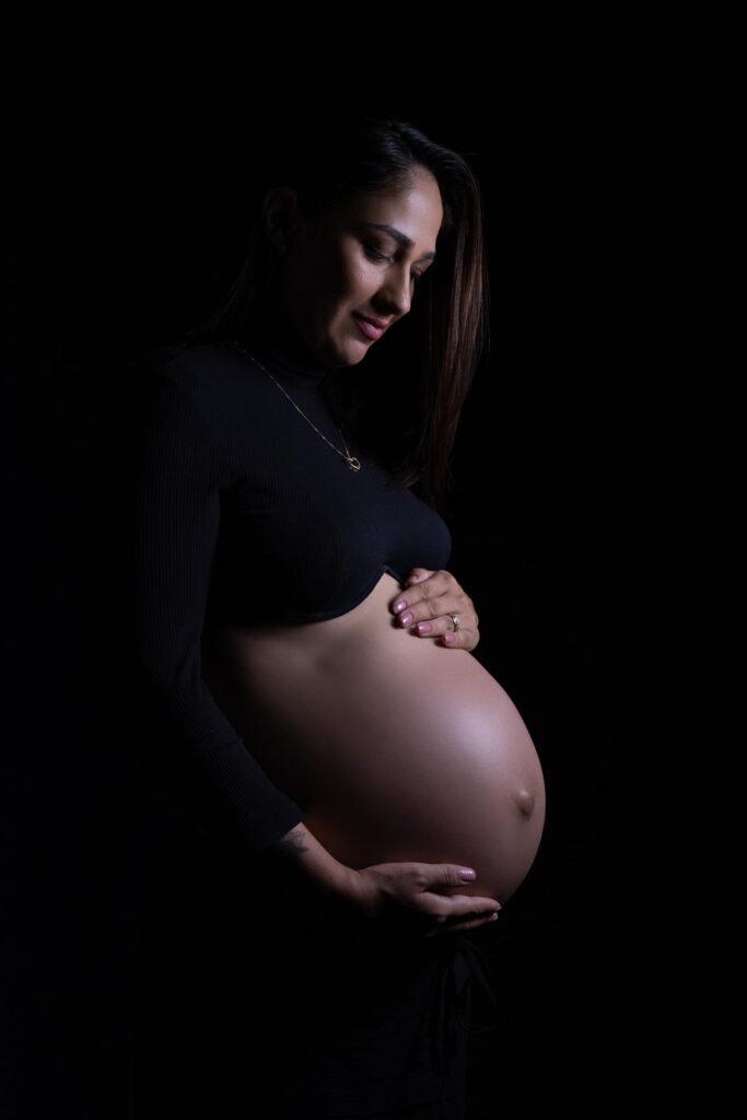 Gestante acariciando a barriga em retrato de estúdio com iluminação lateral e fundo preto.