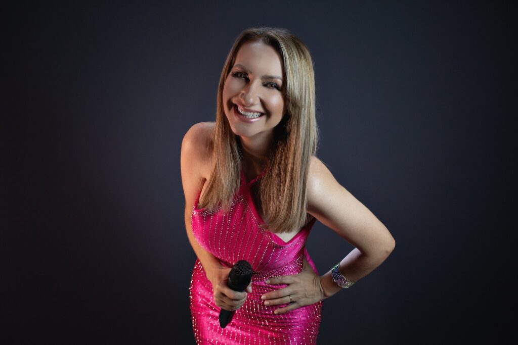 Artista sorrindo com vestido rosa e microfone, em retrato de estúdio sobre fundo escuro.