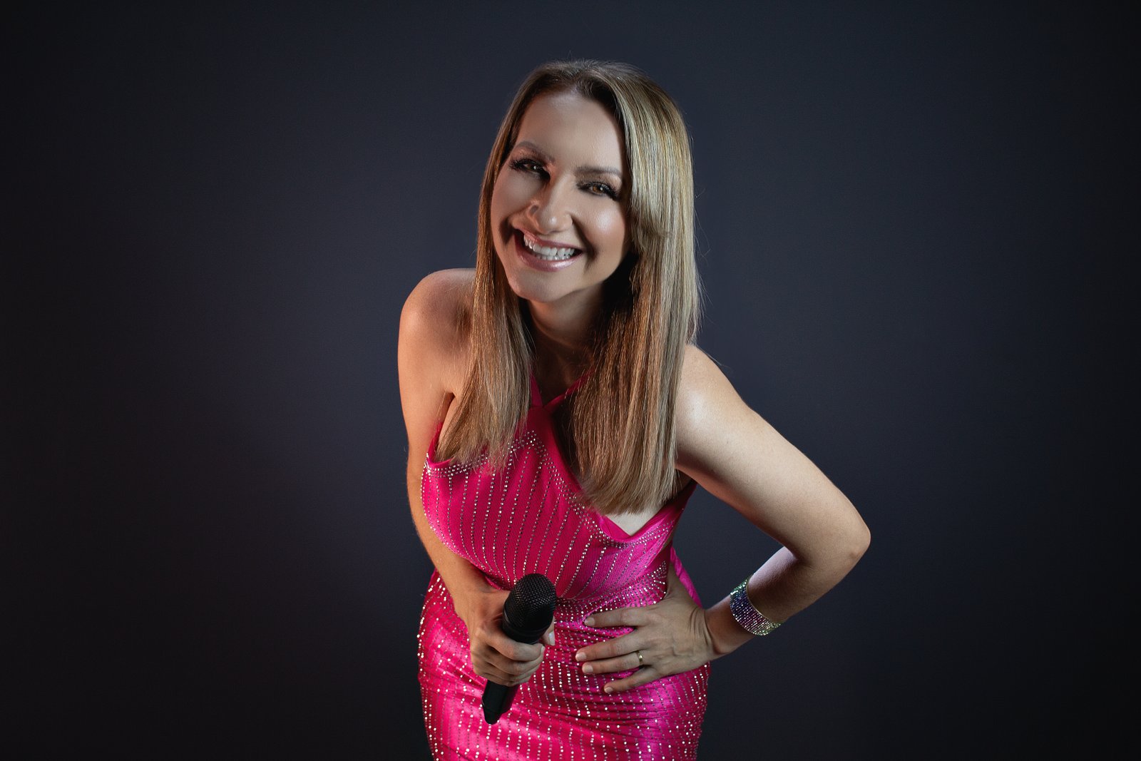 Artista sorrindo com vestido rosa e microfone, em retrato de estúdio sobre fundo escuro.