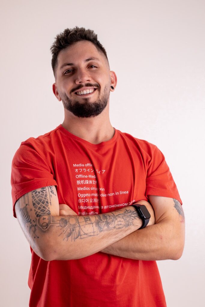 Diego Barbosa sorrindo de braços cruzados, com camiseta vermelha.